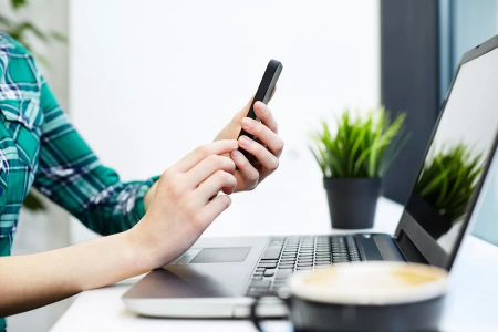 Person using smartphone while working on laptop, surrounded by a plant and coffee cup, representing modern remote communication and productivity in tax relief consultations.