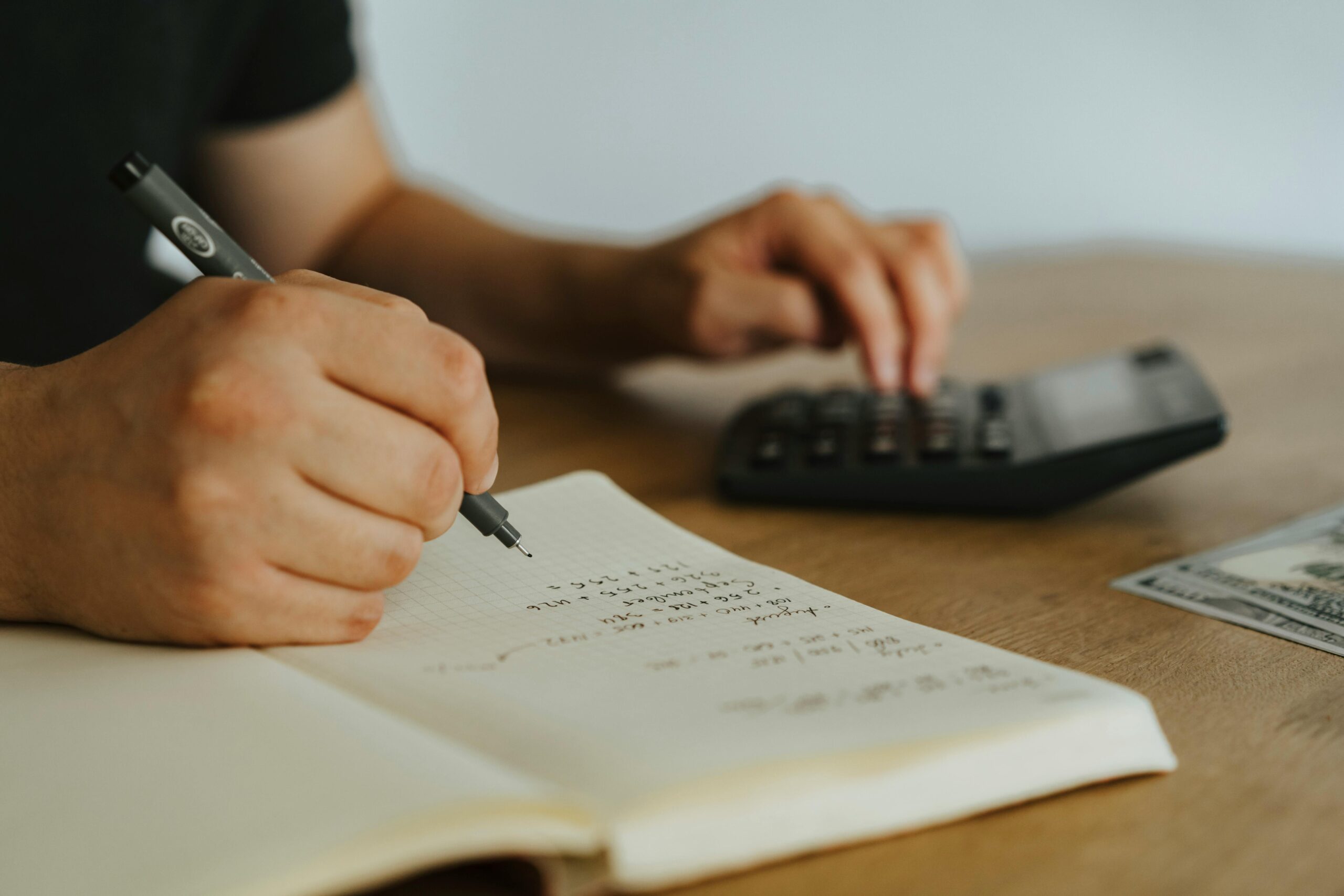 Person writing in notebook with calculator and cash on wooden table, illustrating tax preparation and financial planning.